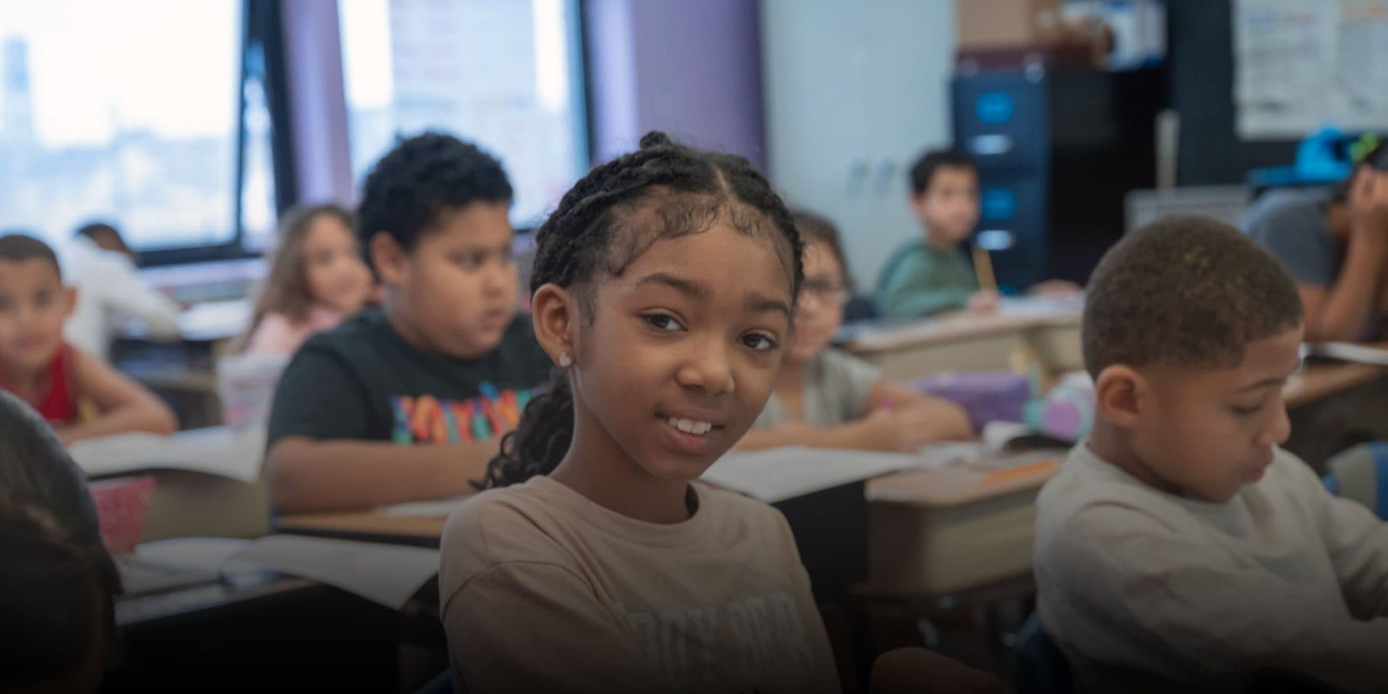 Smiling student at a desk in a busy classroom.