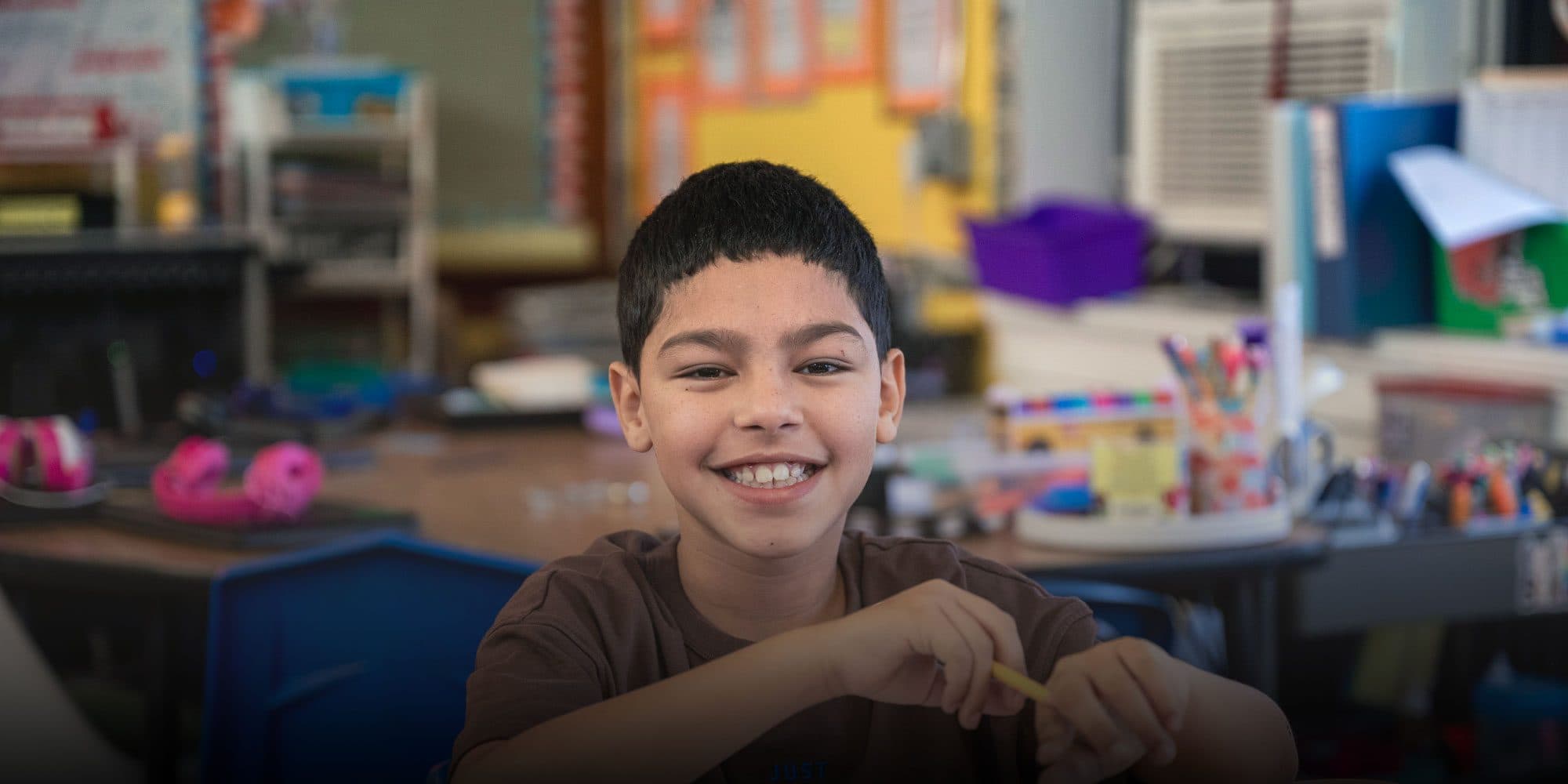 Smiling student holding a pencil at a classroom desk.